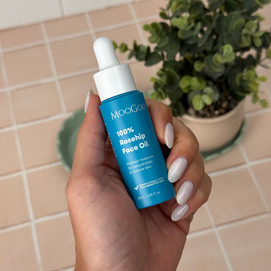 A hand with white manicured nails holding a blue bottle of MooGoo 100% Rosehip Face Oil with a white dropper lid, against a soft pink tiled background with a green potted plant.