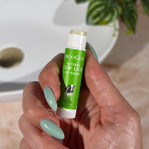 Hand holding a green tube of MooGoo Natural Cow Lick Lip Balm with a plant and bathroom sink in the background.
