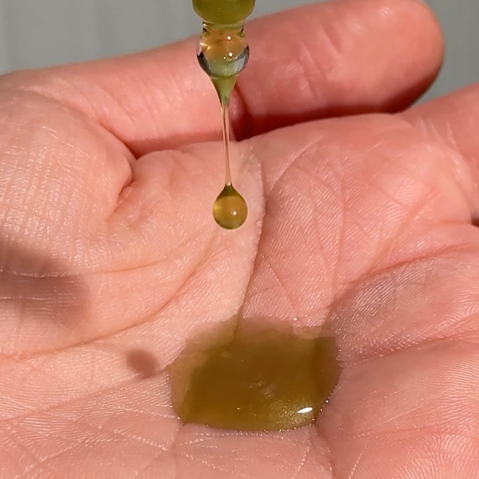 Hand holding a dropper with a small amount of yellow/green liquid, dripping onto the palm.