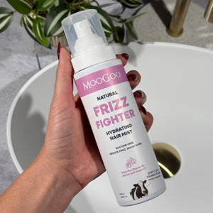 Close-up of hand holding MooGoo Natural Frizz Fighter Hair Mist with purple label above sink, natural hair treatment spray for reducing frizz