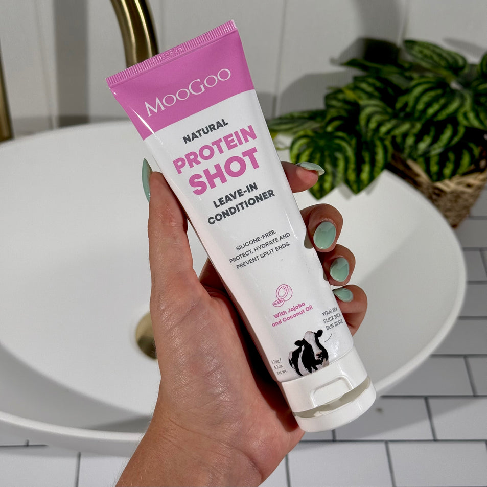 Hand holding a tube of MooGoo Protein Shot Leave-In Conditioner with a plant in the background.