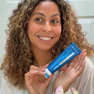 A smiling woman with curly brown and blonde hair holds a blue tube of MooGoo Pepeprmint Toothpaste in front of her face. The toothpaste packaging is blue with white text and a white stripe at the bottom.