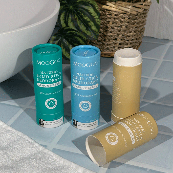 MooGoo Natural Solid Stick Deodorants, Lemon Myrtle, Coconut Cream and Oats & Honey, on a bathroom counter, with a plant in a basket and bathroom sink in the background.