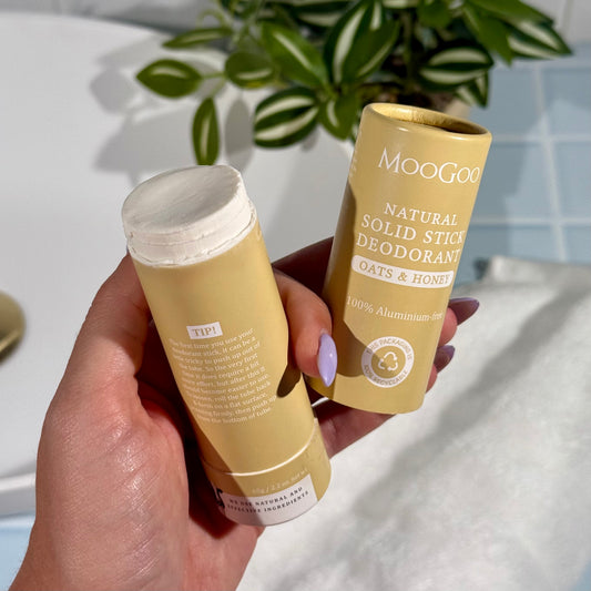 MooGoo Oats & Honey Natural Solid Stick Deodorants held in a hand open and pushed up with a plant in the background.